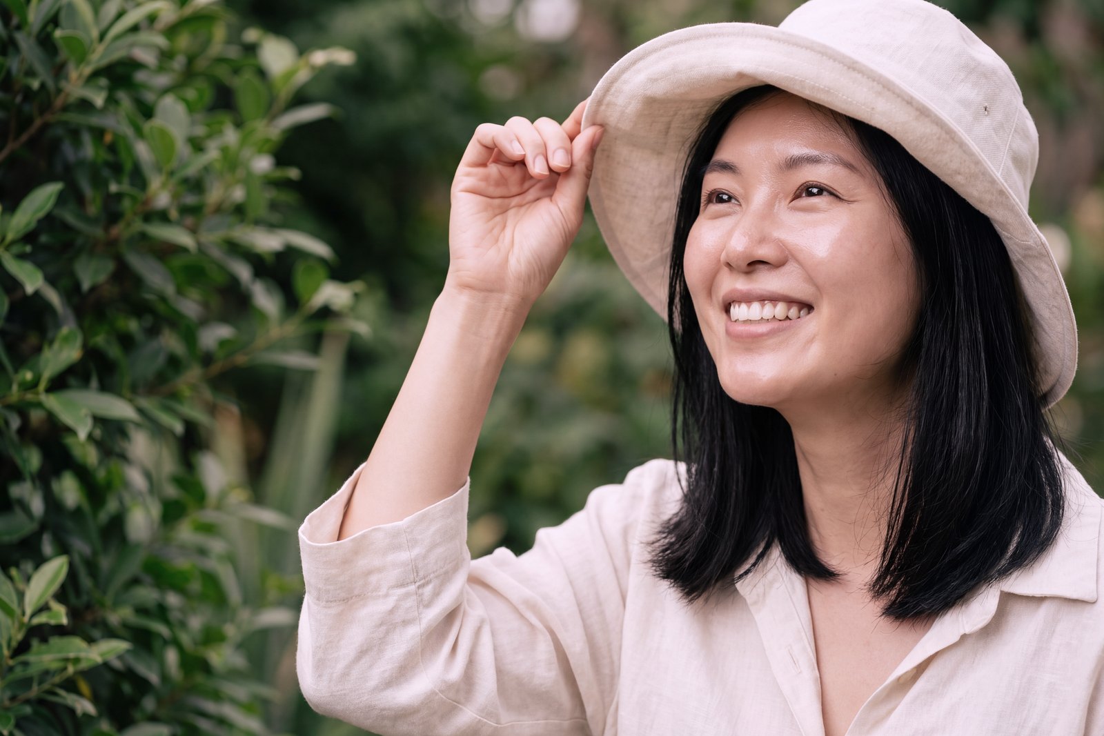 A woman lifting a linen sun hat and smiling up at the sun in a green garden.